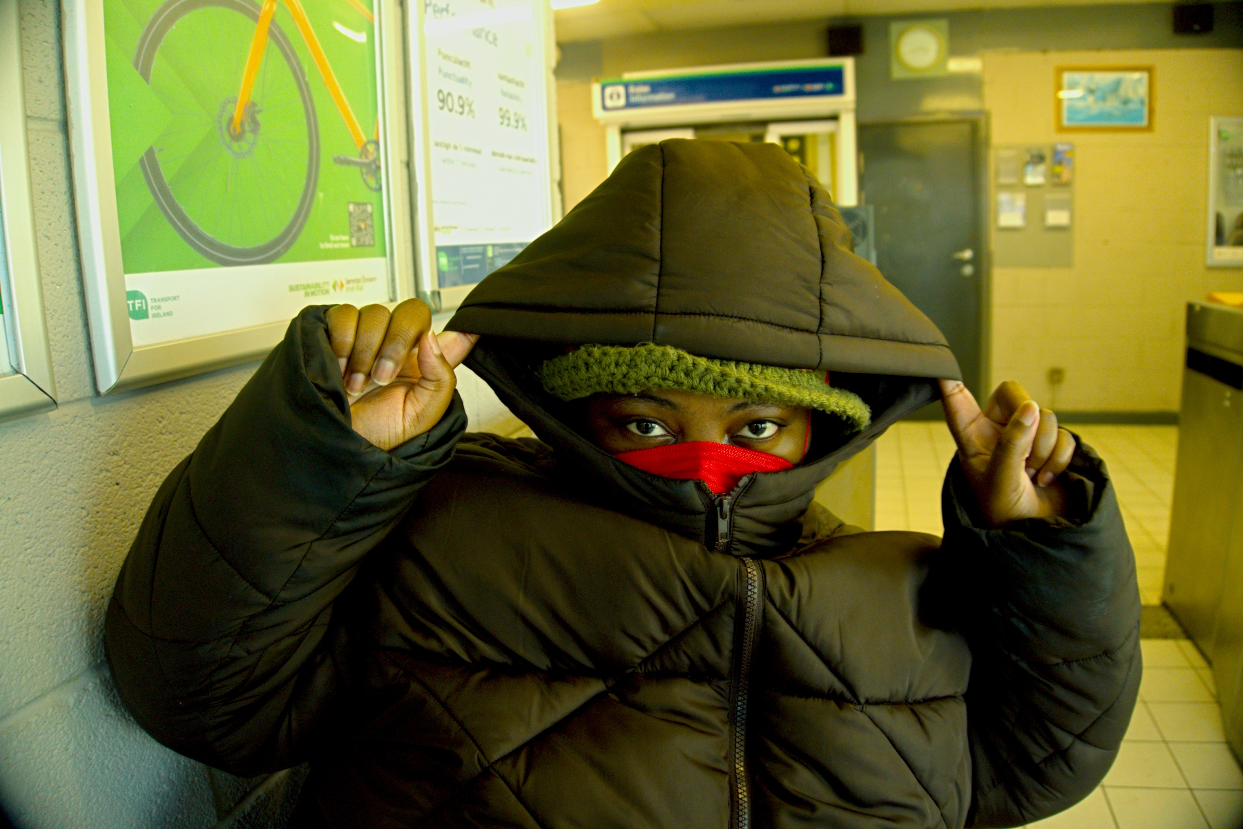 Artist Love, Builder of Worlds in a train station wearing a brown hoodie, red mask and khaki hat. He has his hood up and his fingers stretching the hoodie.